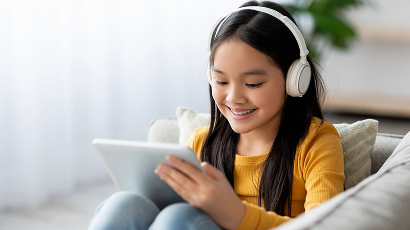 A young girl relaxing at home wearing headphones plays a game on her tablet