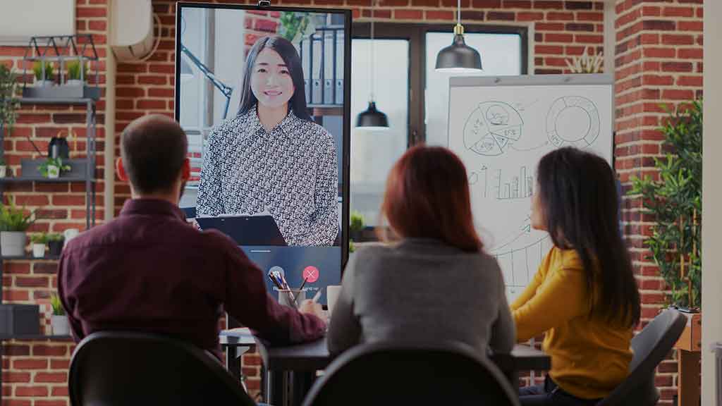 Three employees in a small meeting room on a video conference with a remote worker.