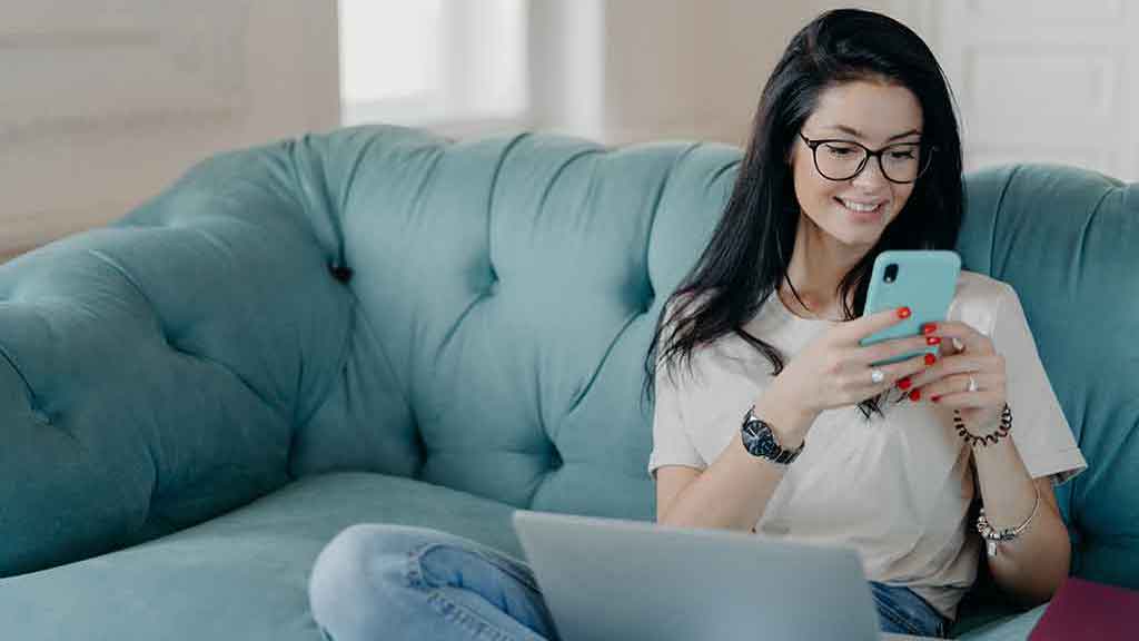 Young woman at home sitting on a comfortable blue sofa with a laptop and using her mobile phone