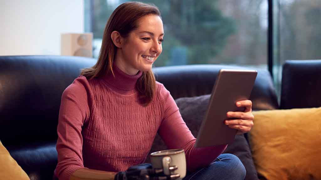 Une jeune femme avec une prothèse de bras se détend à la maison avec une tasse de café en lisant quelque chose sur sa tablette