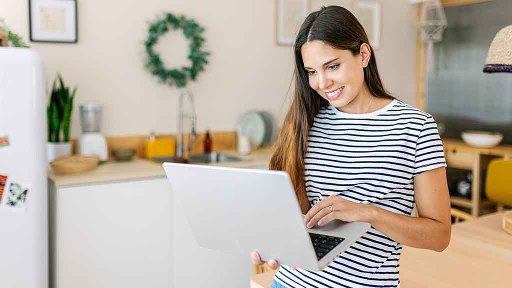 A woman standing in her kitchen holding her laptop as she looks something up online